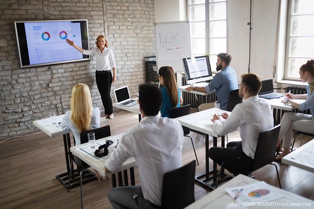 Frau pr&auml;sentiert Diagramme auf Bildschirm vor Seminargruppe in modernem Schulungsraum.