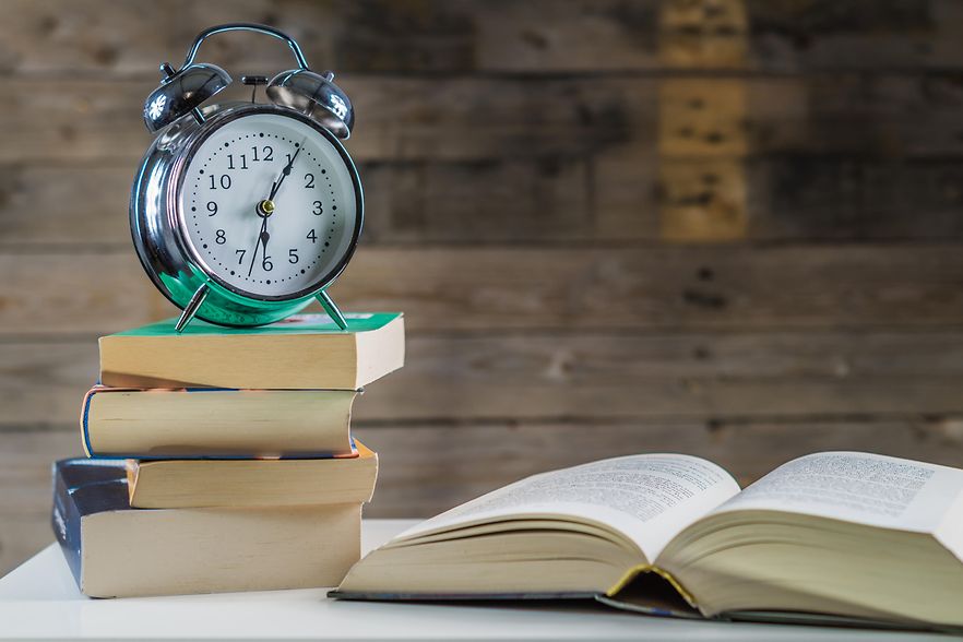 B&uuml;cherstapel mit Wecker darauf sowie aufgeschlagenem Buch