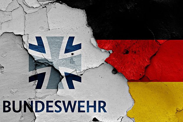 Bundeswehr-Logo auf br&uuml;chiger Wand mit deutscher Flagge im Hintergrund.
