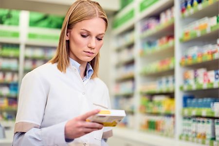 Frau in Apotheker-Kleidung steht in Apotheke, schaut auf Medikamenten-Schachtel