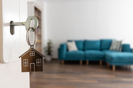Schl&uuml;ssel mit Haus-Anh&auml;nger steckt in T&uuml;r, im Hintergrund modernes Wohnzimmer.