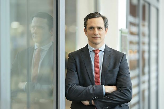 Carl Oetker im Anzug mit roter Krawatte lehnt an Glaswand.