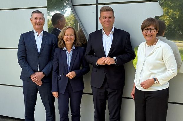 Vier Personen im Anzug und Business-Outfit vor moderner Wand.