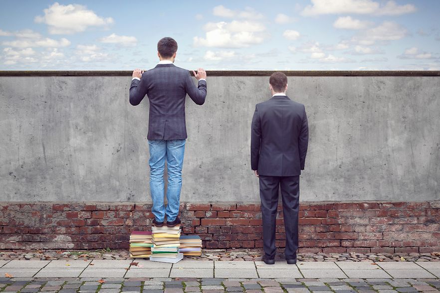 Zwei M&auml;nner vor Mauer, einer schaut mit Hilfe von B&uuml;chern dar&uuml;ber