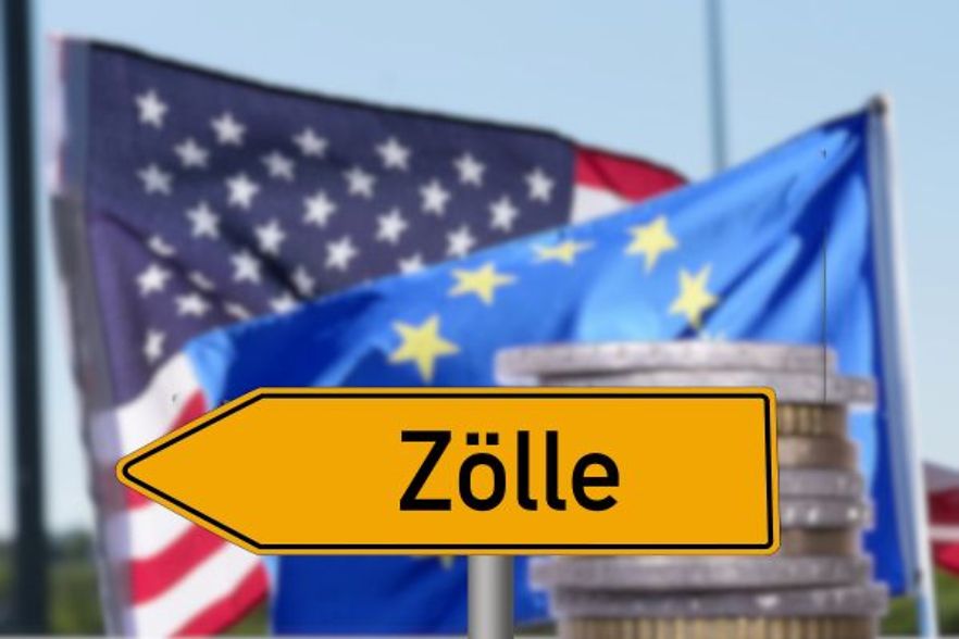 Schild mit Aufschrift &bdquo;Z&ouml;lle&ldquo; vor Flaggen der USA und EU sowie M&uuml;nzstapeln