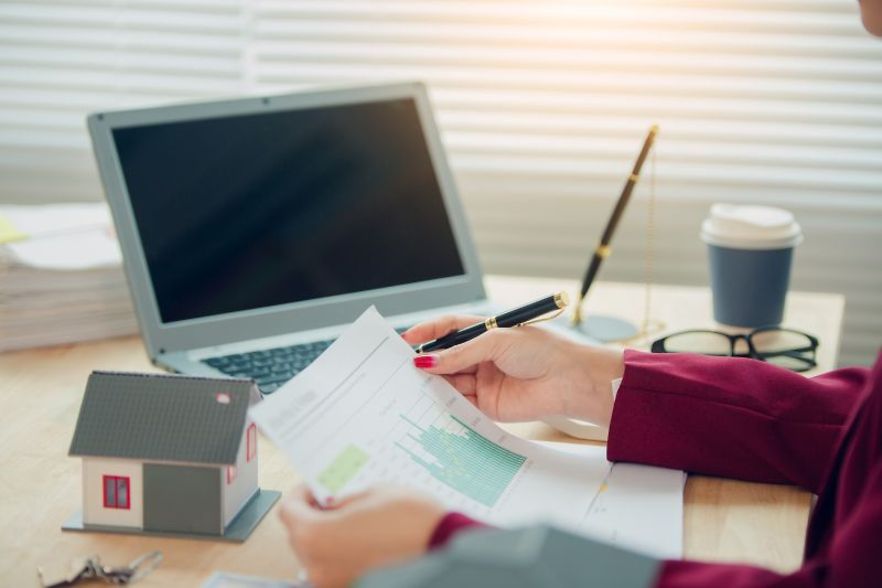 Symbolfoto einer Person, die Finanzdokumente vor einem Laptop pr&uuml;ft.