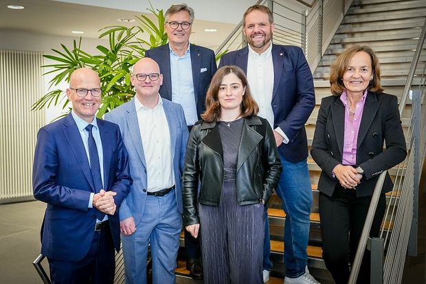Sechs Personen stehen auf einer Treppe und l&auml;cheln in die Kamera, Gruppenfoto in gesch&auml;ftlicher Umgebung.