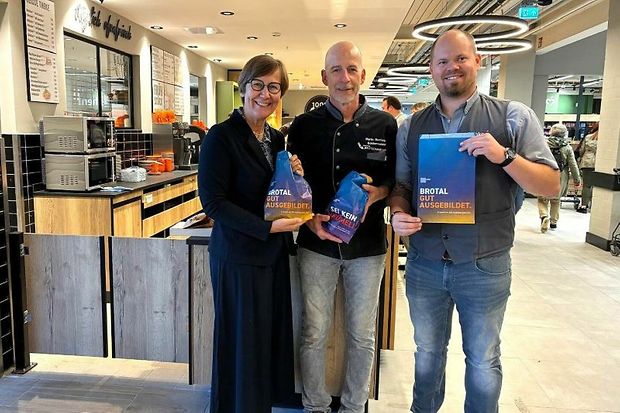 Drei Personen stehen in einer B&auml;ckerei und halten Werbematerial mit der Aufschrift &bdquo;Brotal gut ausgebildet&ldquo; in den H&auml;nden.