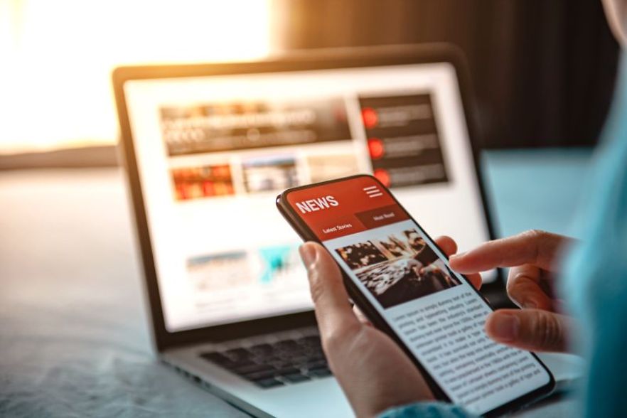Person liest Nachrichten auf dem Smartphone vor einem ge&ouml;ffneten Laptop.