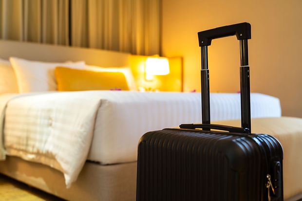 Koffer steht in einem gem&uuml;tlich beleuchteten Hotelzimmer vor einem frisch gemachten Bett