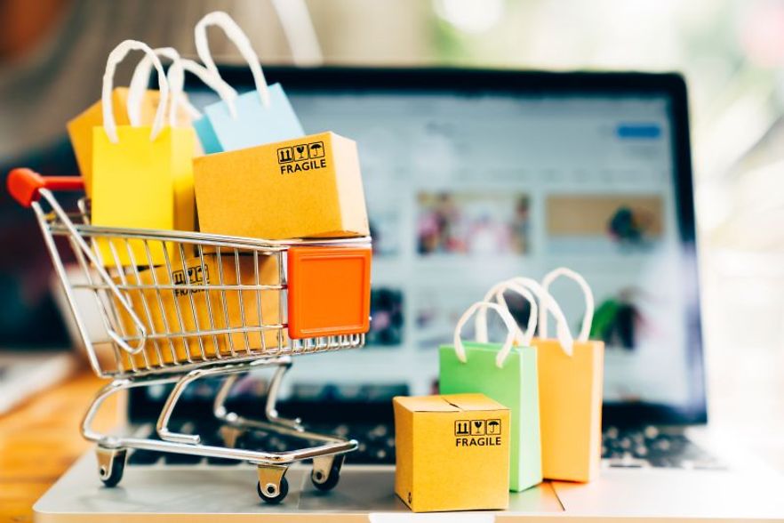 Kleiner Einkaufswagen mit bunten T&uuml;ten und Paketen steht vor einem ge&ouml;ffneten Laptop als Symbol f&uuml;r Online-Shopping.