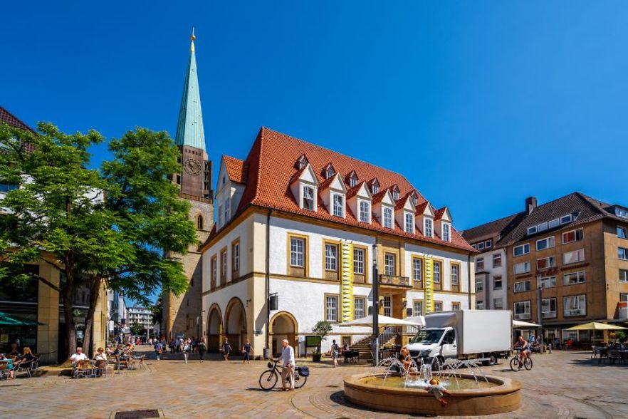Marktplatz in der Bielefelder Altstadt mit historischem Geb&auml;ude, Brunnen und Kirchturm bei sonnigem Wetter.