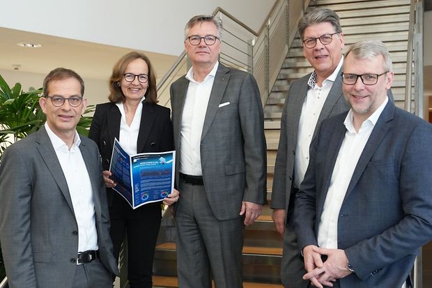 F&uuml;nf Personen im Business-Dress stehen vor Treppe, eine h&auml;lt eine Brosch&uuml;re
