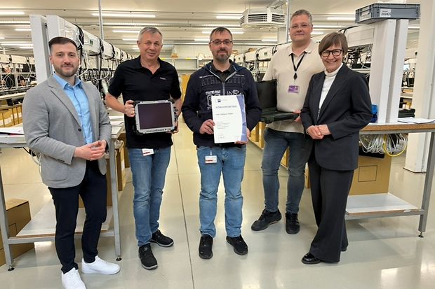 F&uuml;nf Personen in Werkhalle, zwei halten Tablet und Zertifikat